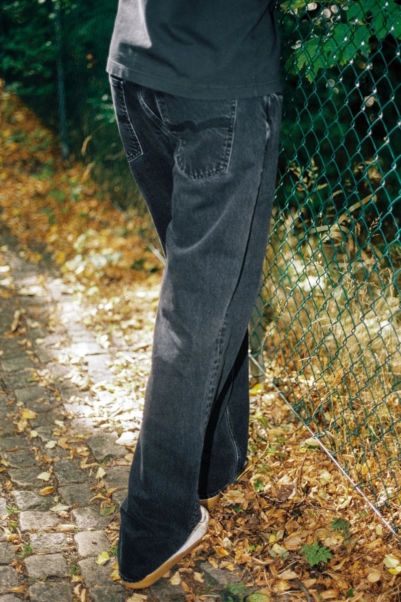 Person wearing dark jeans standing on a leaf-covered ground with a green fence in the background.