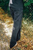 Person wearing dark jeans standing on a leaf-covered ground with a green fence in the background.