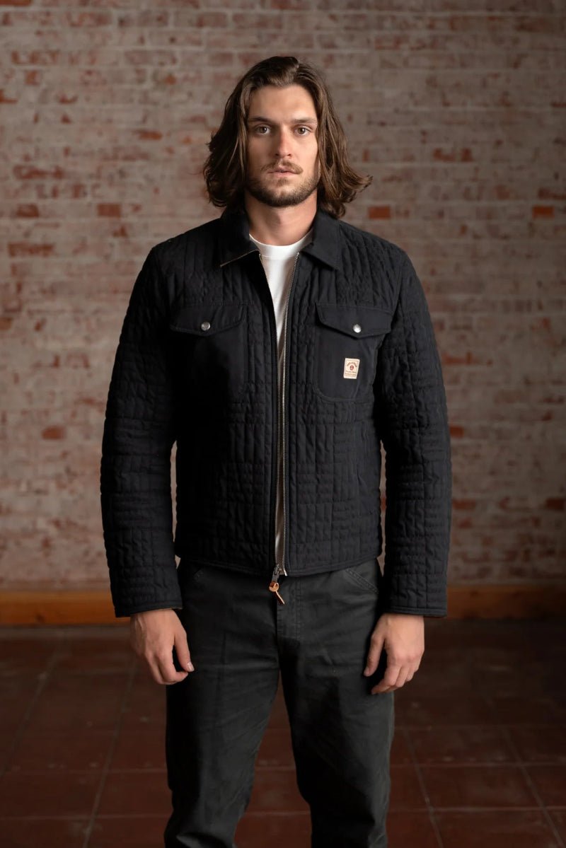 Man wearing a black quilted jacket with a brand logo against a brick wall.