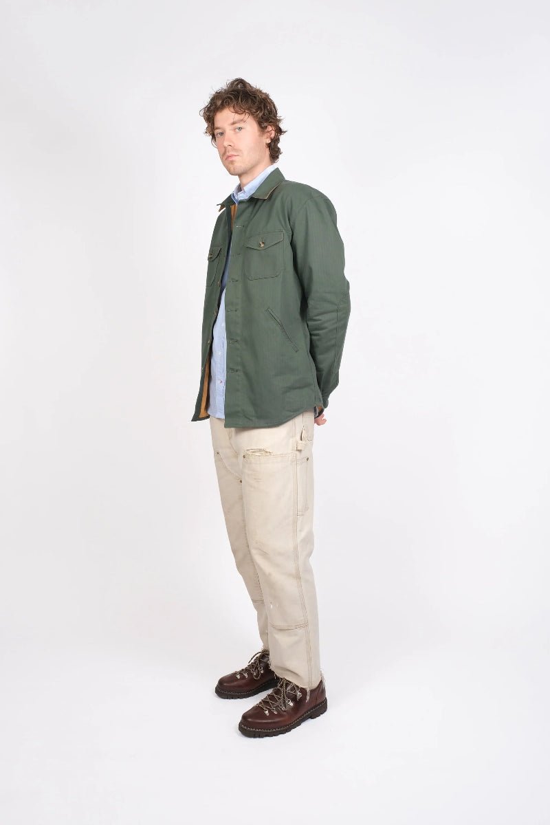 Man wearing a green jacket, light shirt, beige pants, and brown shoes on a white background