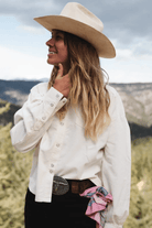 A woman wearing a cream cowboy hat, a cream western-style button-up shirt, and a black skirt with a large silver belt buckle, standing outdoors in a mountain landscape.