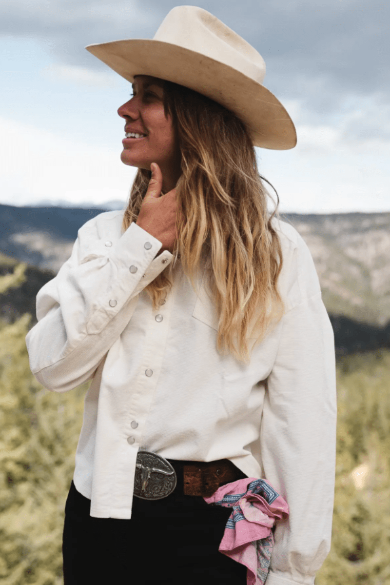A woman wearing a cream cowboy hat, a cream western-style button-up shirt, and a black skirt with a large silver belt buckle, standing outdoors in a mountain landscape.