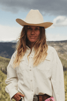 A woman wearing a cream-colored cowboy hat and matching long-sleeve western shirt, standing outdoors in front of a blurred mountain landscape.